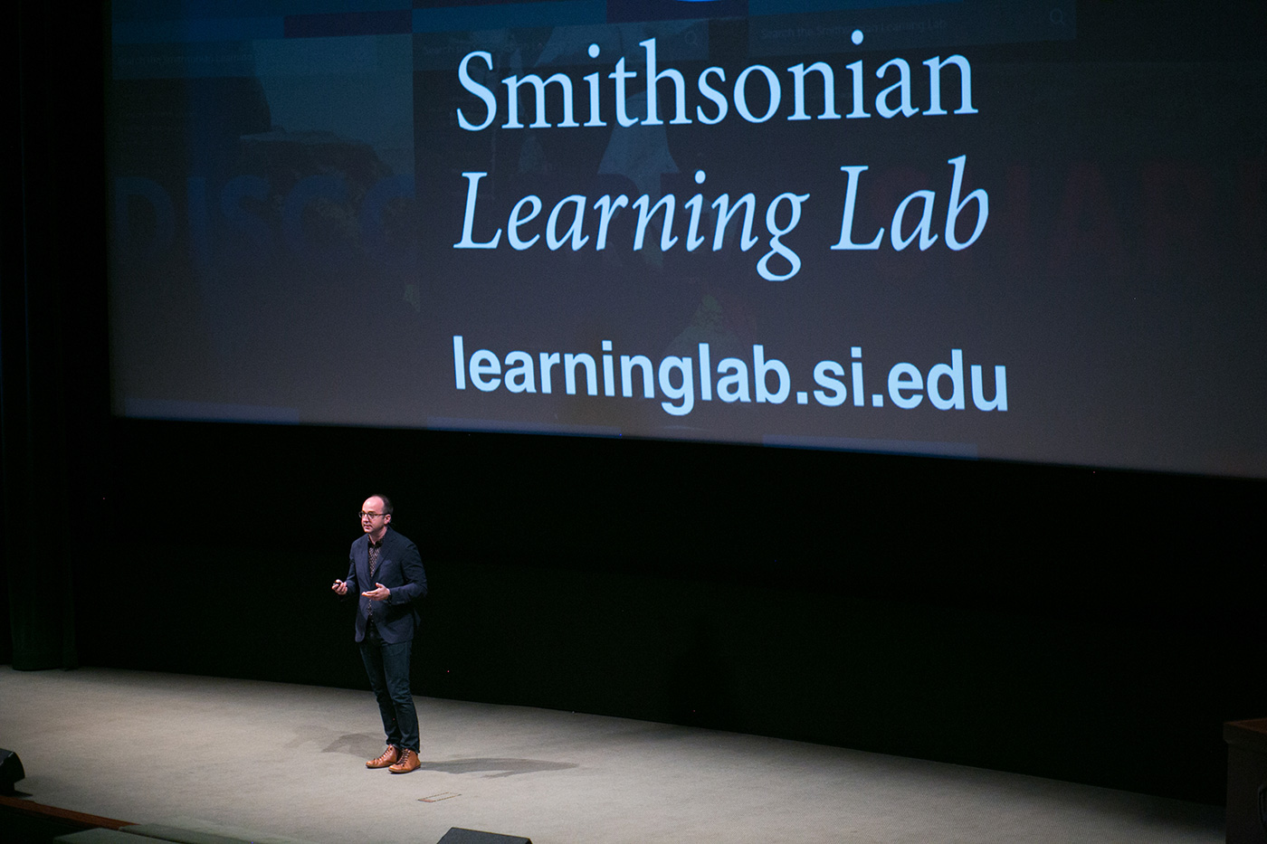Darren Milligan on a large stage presenting the Learning Lab. The screen behind him shows the name and URL of the site.