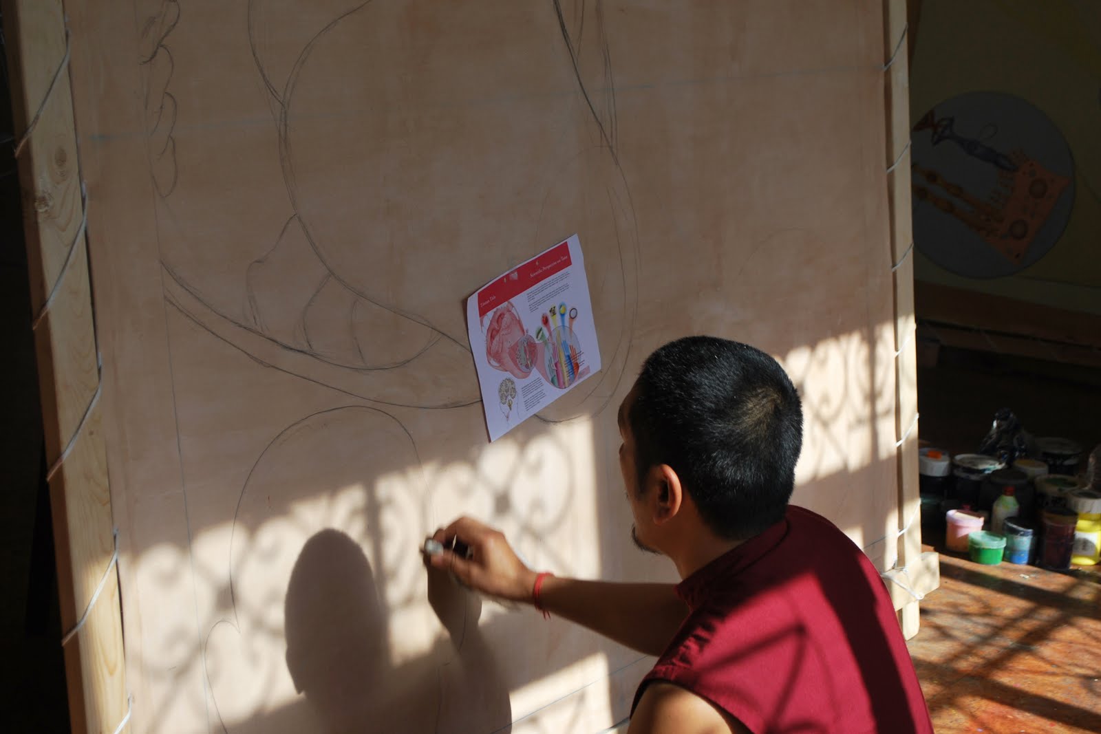 A thangka artist sits in front of a large canvas with a pencil sketch of a large open mouth.